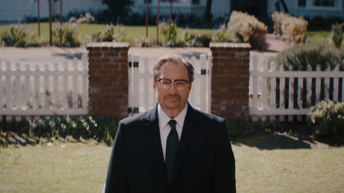 Stephen Root in a suit stands in front of a literal white picket fence in Happily