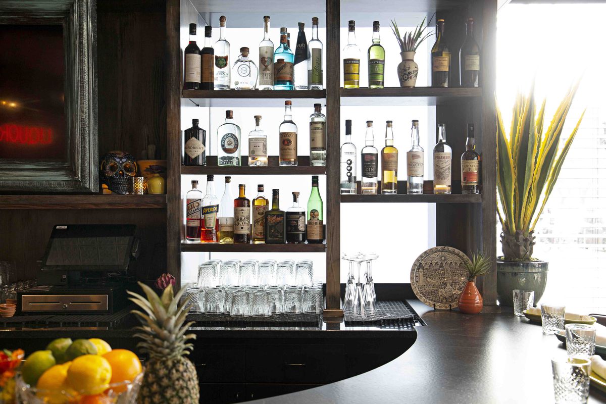 A counter-level view of a restaurant bar with backlit shelves of liquor bottles, bowls of citrus, and place settings