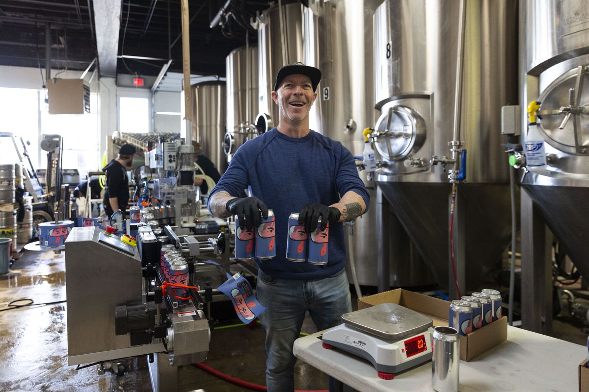 Darren Provenzano holds two four-packs of beer in front of large stainless steel brewing equipment at Fracture Brewing.