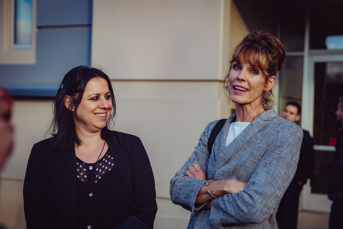 Two women dressed in business attire smile and appear relaxed, outside a concrete-walled building.