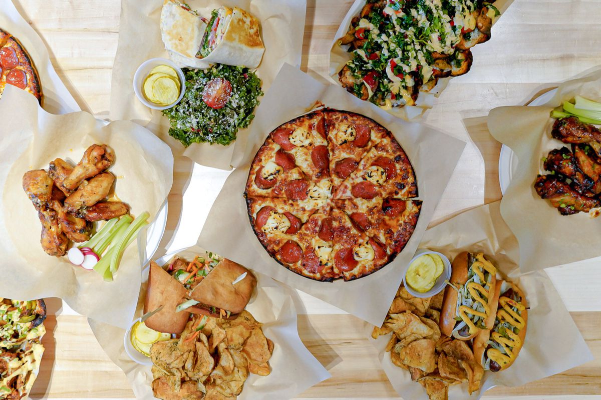 A crispy-edged pizza surrounded by other bar snacks and drinks on a wooden table.