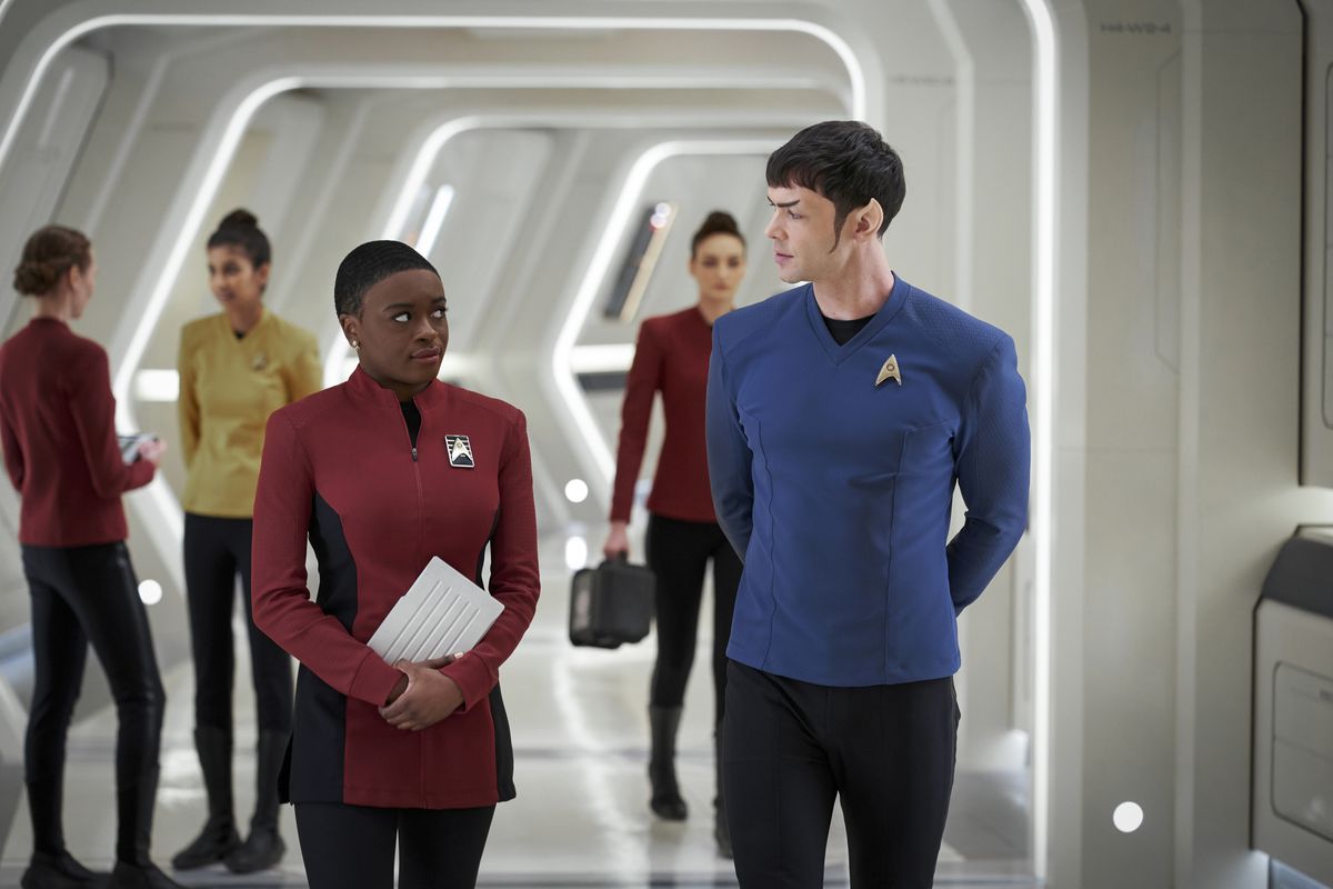 Uhura and Spock walking in the halls of the Enterprise; she is holding a tablet in front of her and looking at him, he is walking with his arms behind his back looking at her. There are other Enterprise personnel in the background
