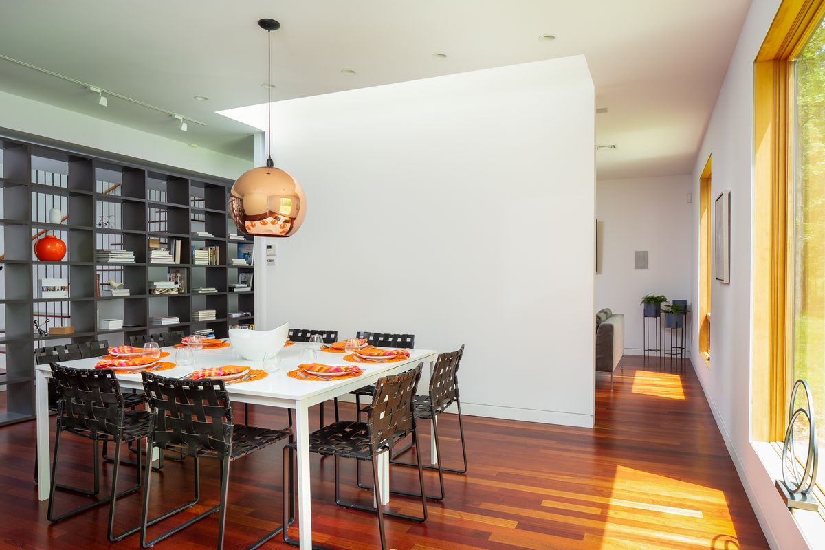A white square dining room table with eight black chairs, wood floors, and a copper pendant lamp.