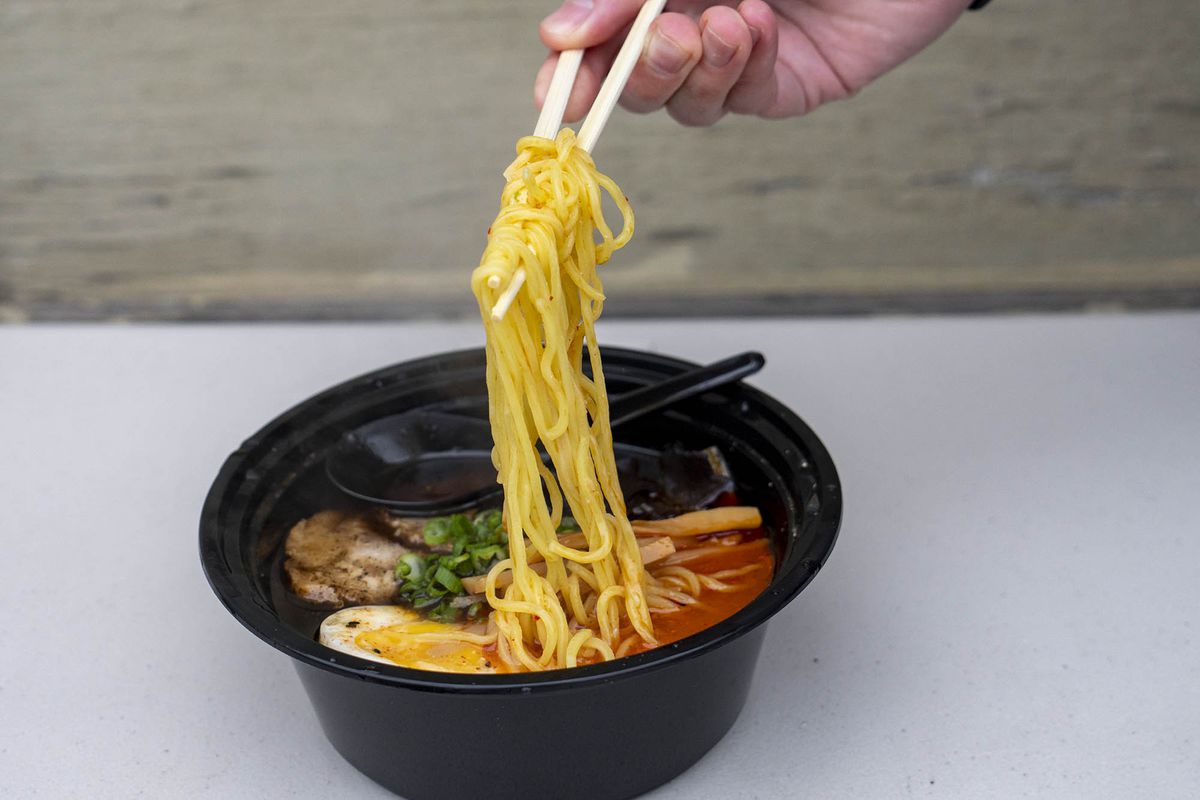 A person pulls noodles from a takeout black ramen bowl with wooden chopsticks.