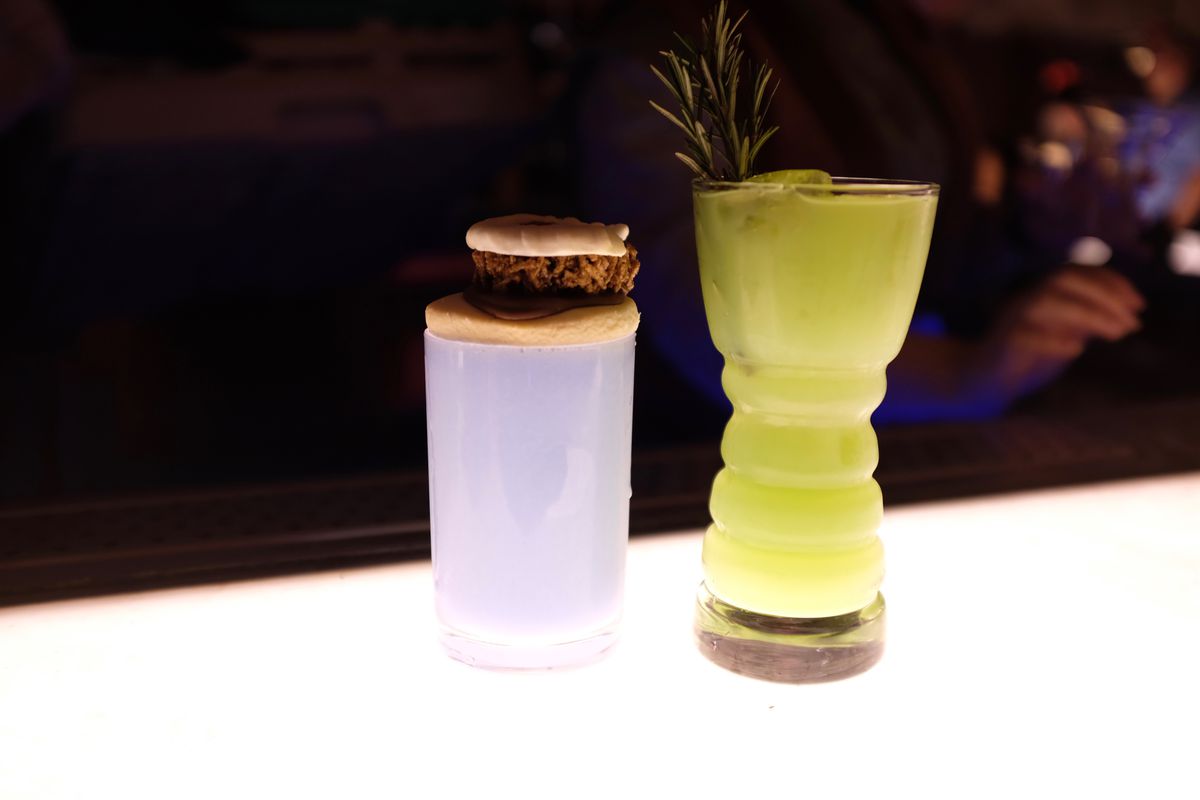 Two bright cocktails in decorative glassware, one topped with a cookie sandwich, on a bright, back-lit bar