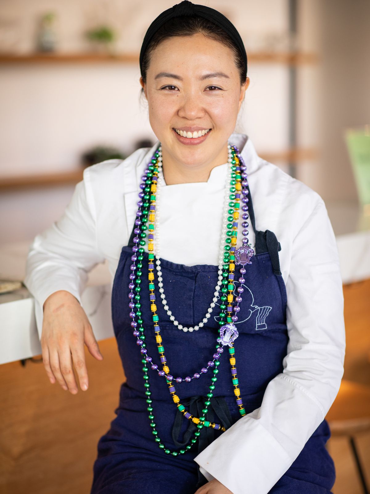 A woman smiling at the camera wearing Mardi Gras beads, a blue apron, and a white chef’s coat