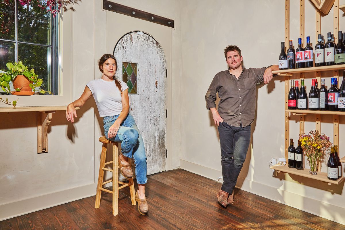 A white woman in jeans leaning against a stool and a white man in jeans leaning against a wall of wine. 