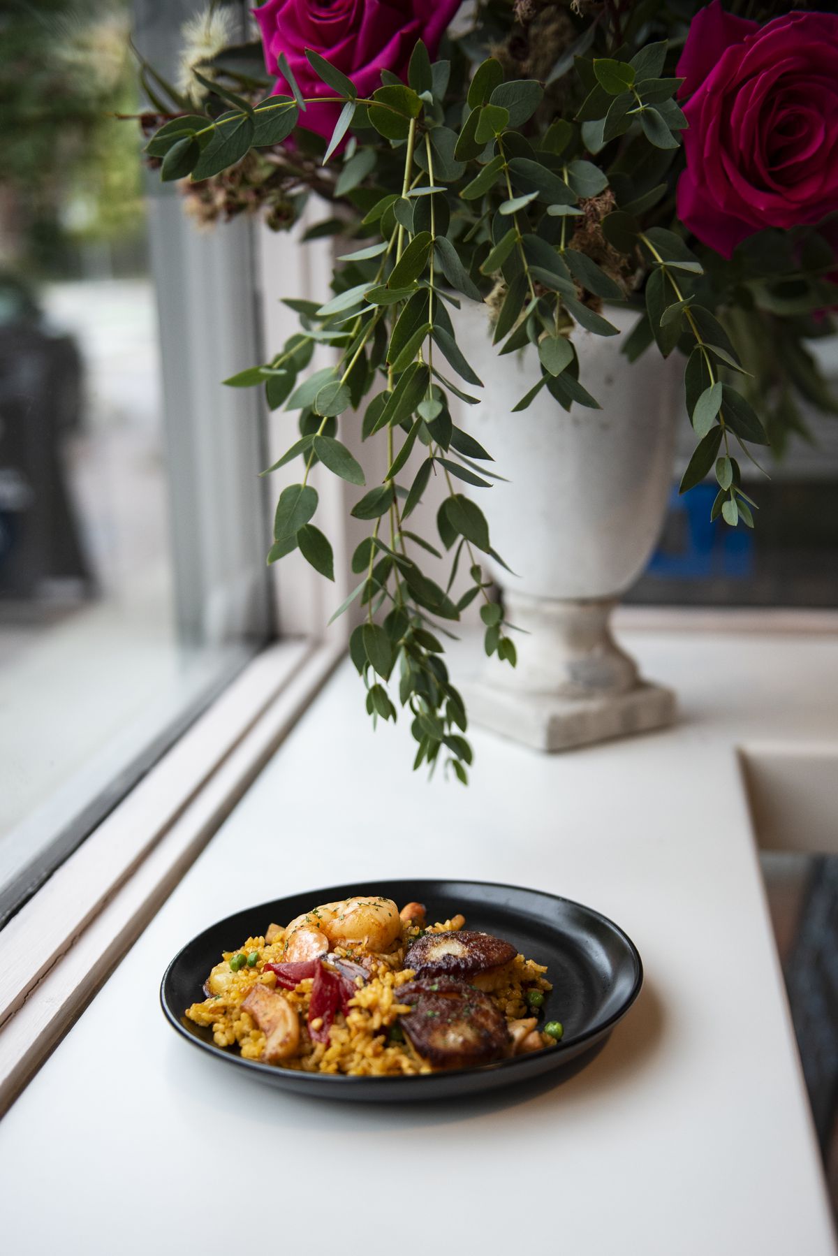 A plated dish of paella next to a white vase of roses on a white surface next to a window.