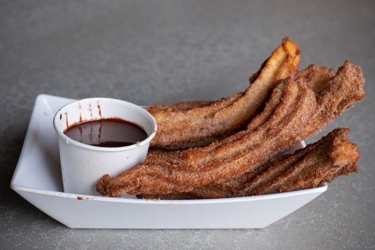 A white boat holds several deep fried and sugar coated churros and a cup of dark, liquid chocolate