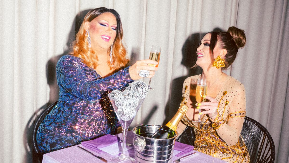 Two drag performers sit at a table and toast with Champagne glasses