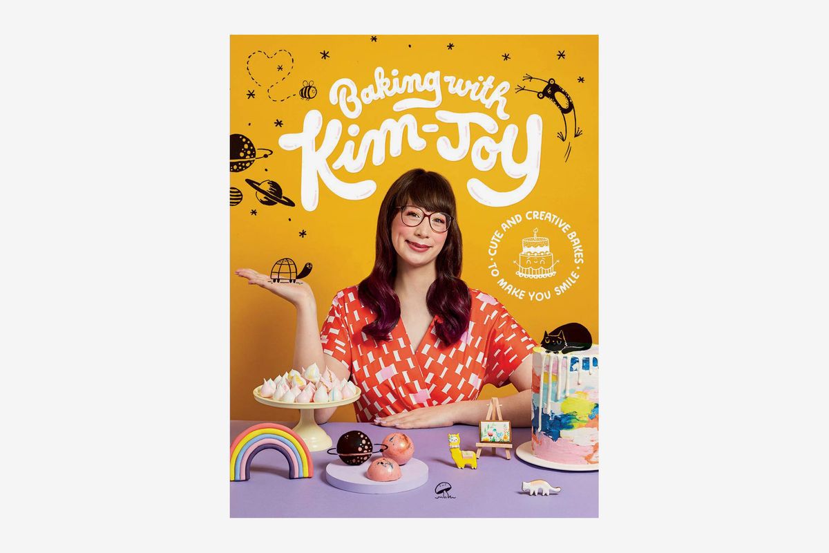 The cover of “Baking with Kim-Joy” featuring Kim Joy posing with a table full of baked goods