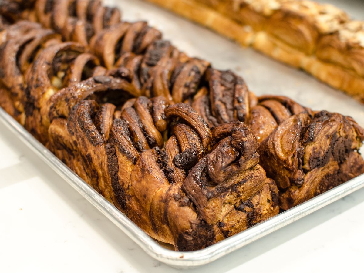 Two long, narrow loaves of chocolate babka sit side by side on a silver tray, cutting diagonally across the front of the photo. In the background, almond babka is a bit visible.