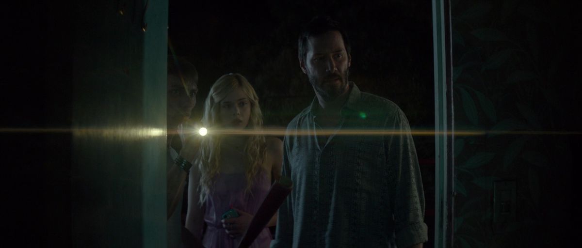 Keanu Reeves and Elle Fanning peer inside a room in The Neon Demon as a flashlight shines next to them.