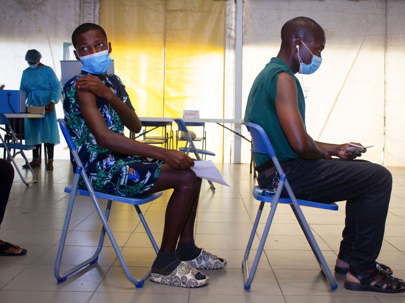 Two people wearing breathing masks sitting in folding chairs in a line in a clinic.