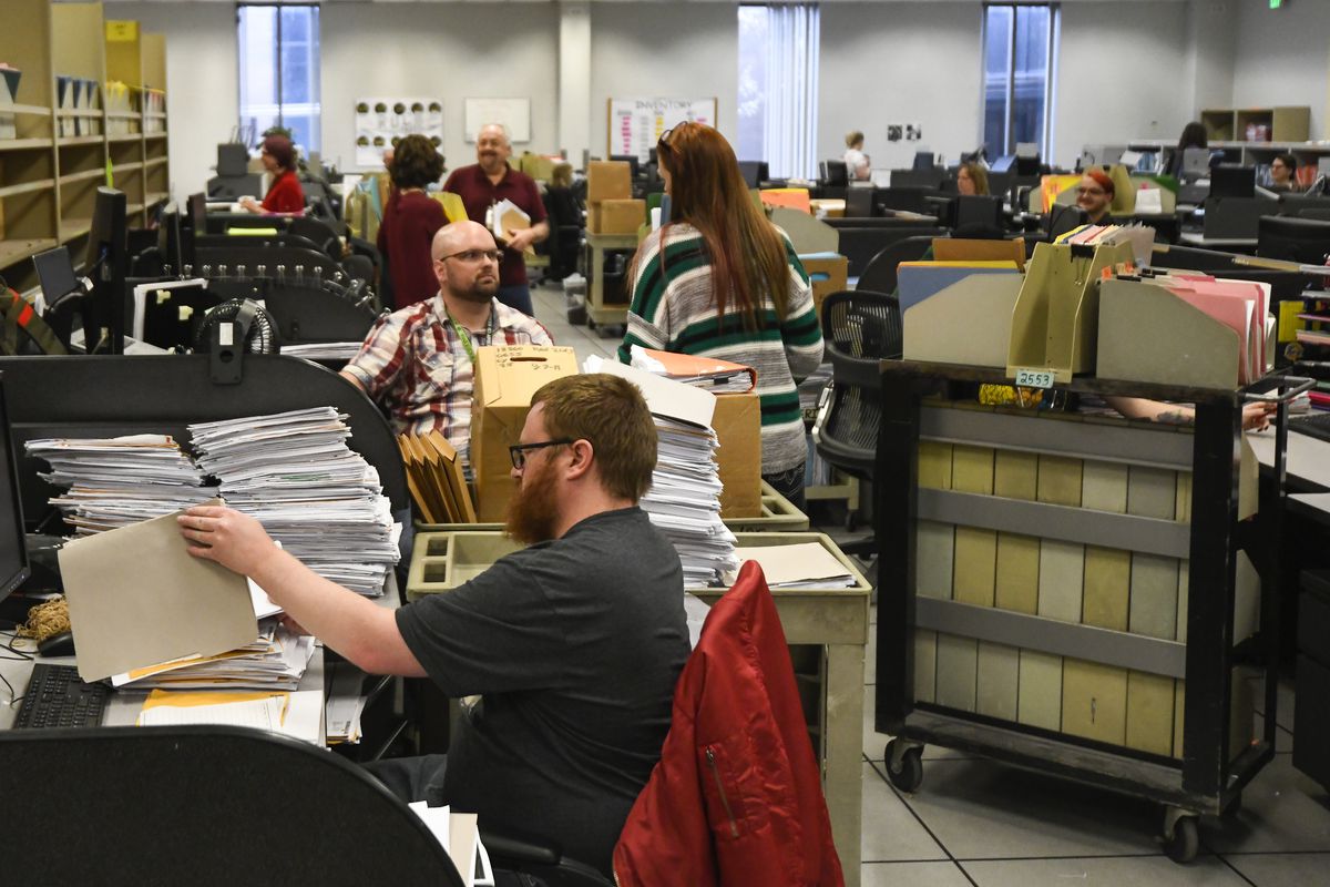 Employees work at desks in a room crowded with stacks of paper files.