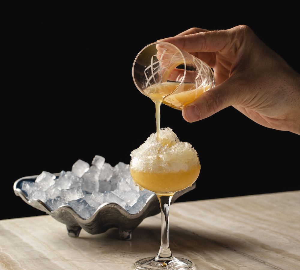 A hand pouring an orange cocktail into a coupe glass.