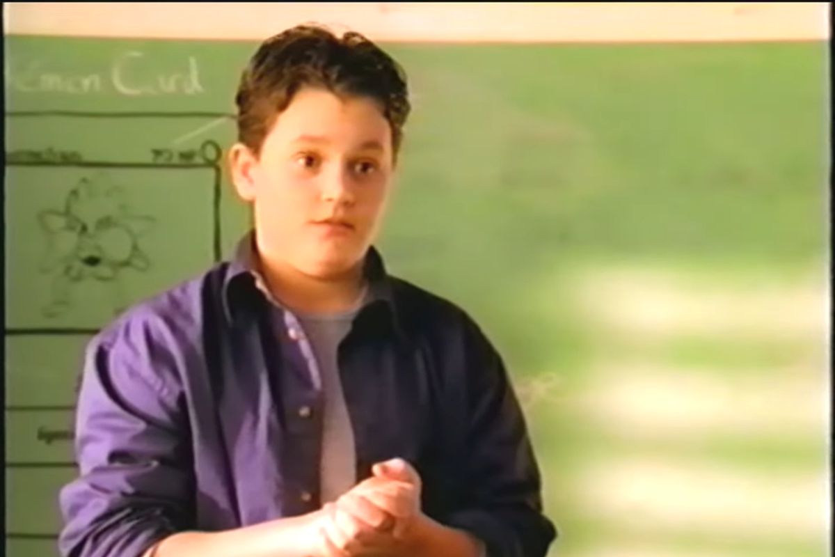 A still from a video showing Penn Badgley standing in front of a chalk board. He’s holding his hands together as he looks out towards the class. 