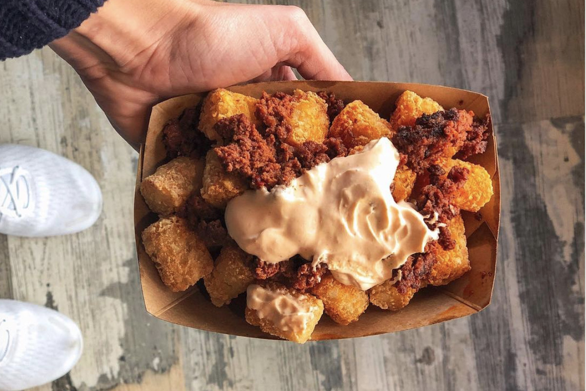 A person with light skin holds a disposable paper container filled with tater tots smothered in red smoked chorizo and creamy sriracha sour cream.