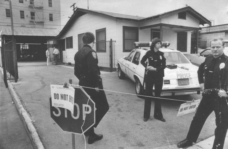 A house. Police officers stand in a driveway with a police car outside of the house. There is a Stop sign. 