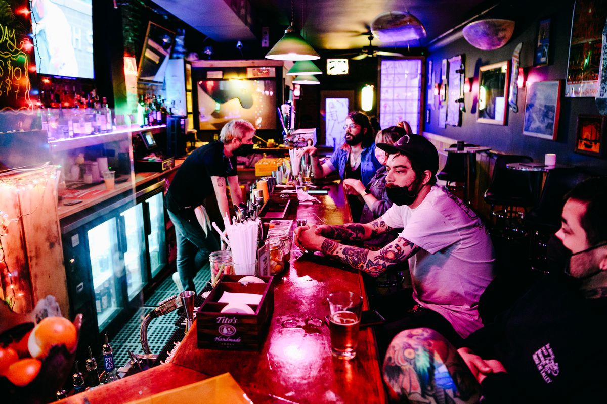 Masked and unmasked customers sit at a bar top in Brooklyn, talking with a bartender and sipping drinks.