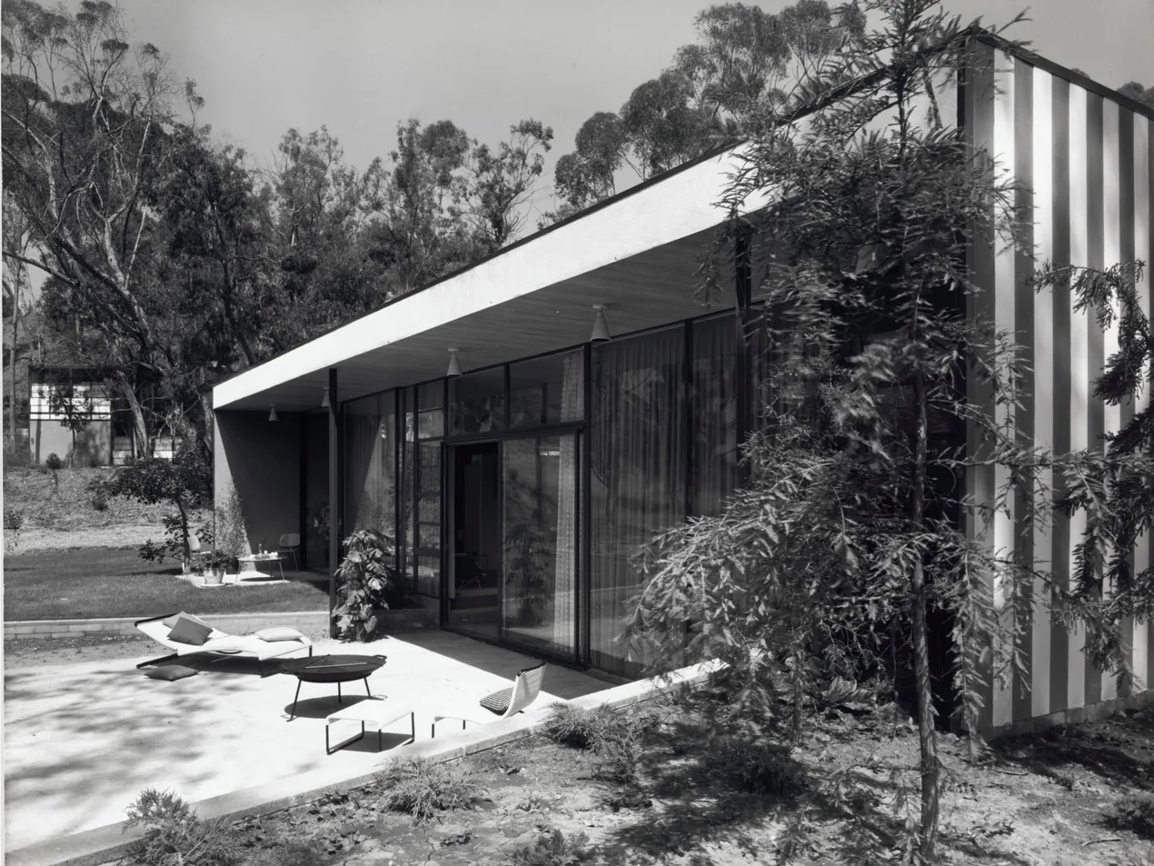 The Entenza House exterior. The roof is flat and the exterior has floor to ceiling windows. There are trees surrounding the house. There is an outdoor seating area.