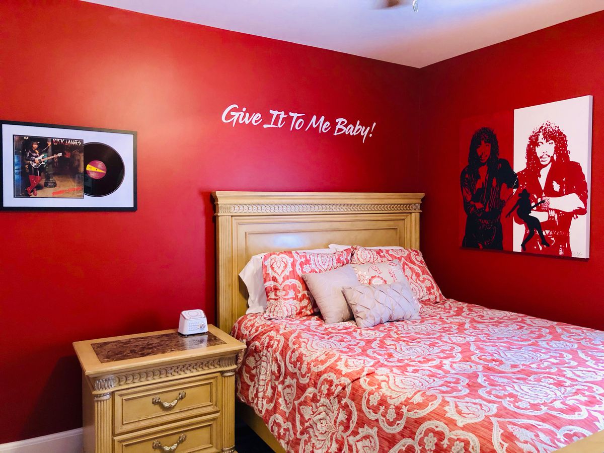 A bedroom with red walls and a screen print of Rick James, and framed 45-inch vinyl. 