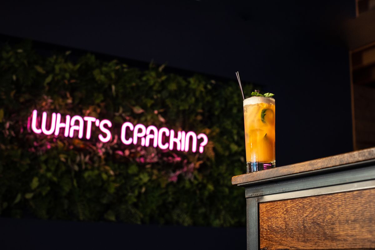 An orange cocktail in a tall glass sits at the edge of a table in front of wall of green plants and a pink neon sign that reads, “What’s Crackin’”