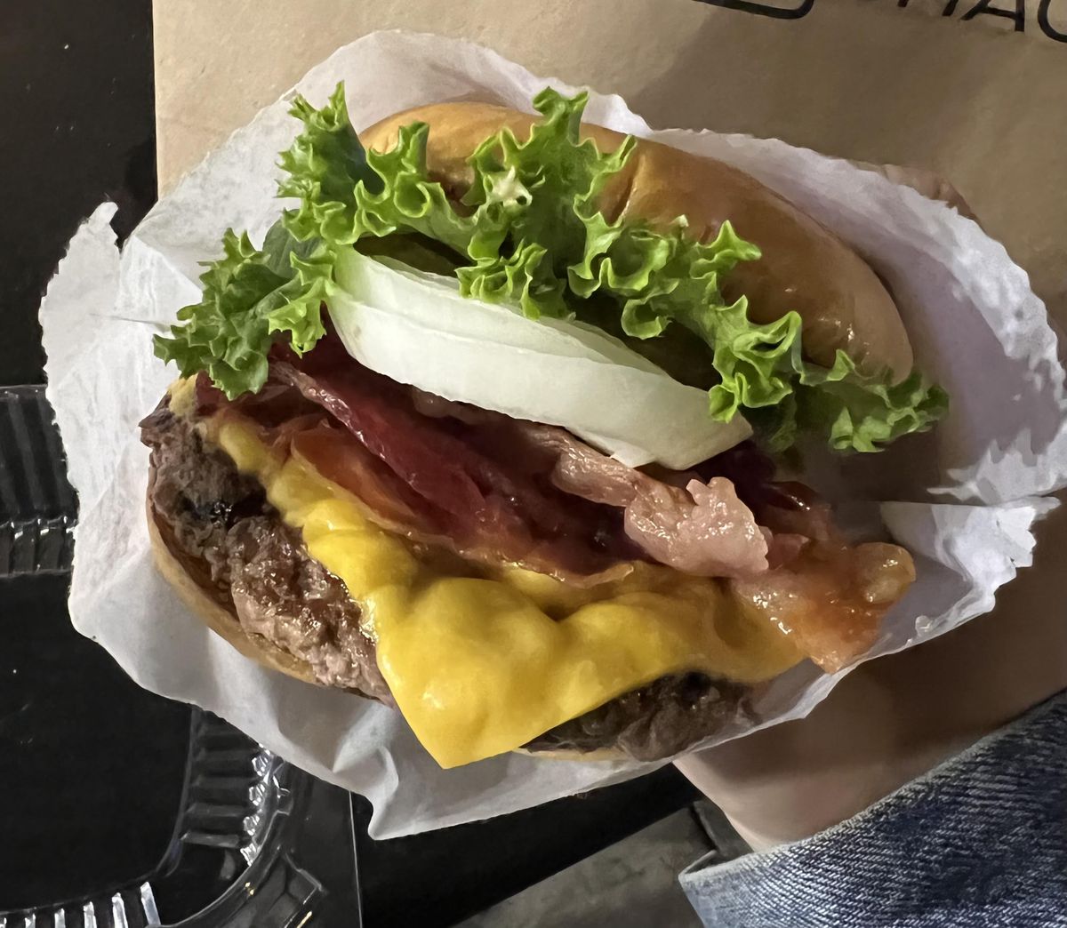 A patron is shown holding a burger in wax paper up to a camera, showing the contents of the fast food snack: white onions, yellow American cheese, bacon, and lettuce
