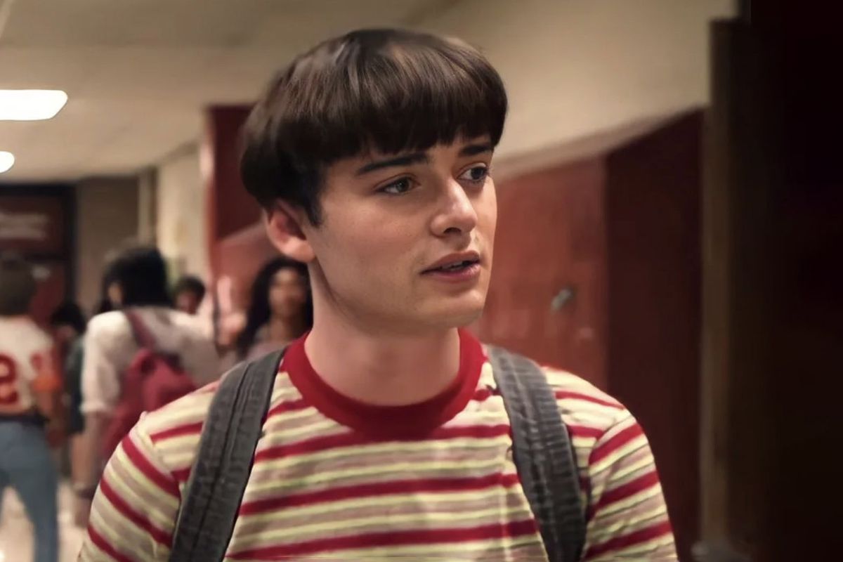 Noah Schnapp as Will Byers, walking through the hall of high school with a bowl cut and a striped red and yellow shirt while wearing a backpack in Stranger Things