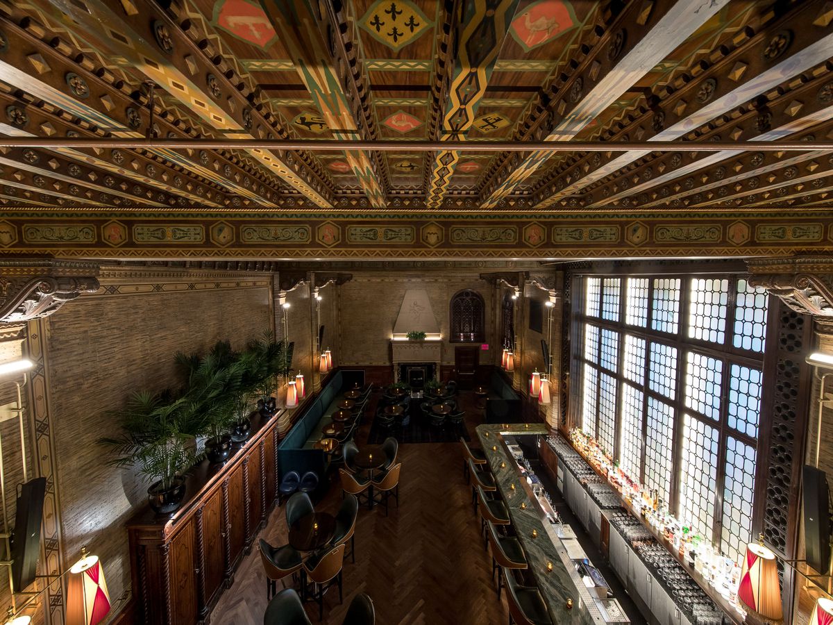 The interior of the Campbell Apartment in Grand Central Terminal, New York City. The wooden ceiling has ornamental decorations. There is a bar with seating. There are many chairs and tables. Tall windows run across one of the walls of the bar.