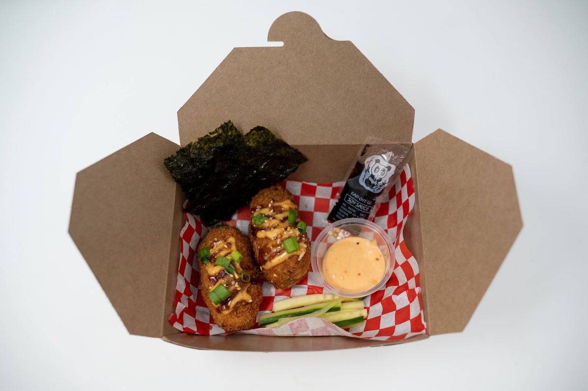 A cardboard to-go package holds two pieces of seaweed wrap, two fried sushi bakes, a plastic container of yum yum sauce, a packet of soy sauce, and slices of cucumber.