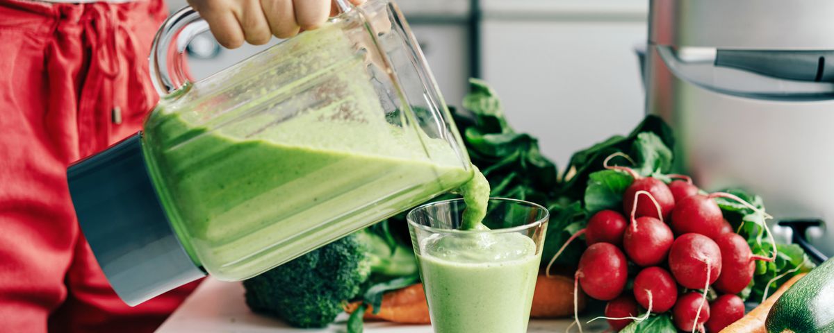 A person pours a green smoothie from a blender into a glass with an array of vegetables like radishes, cucumbers and carrots on the table next to them
