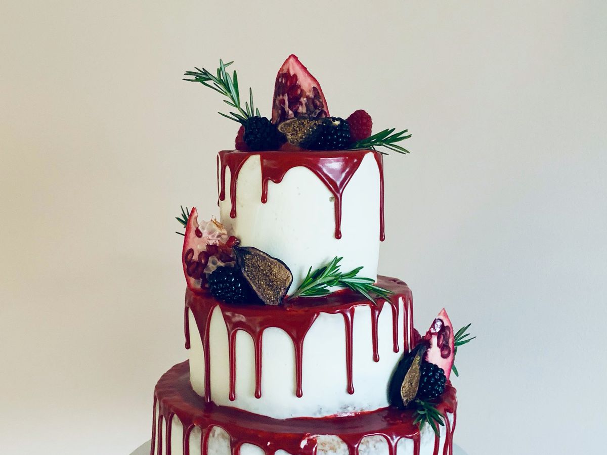 A hand holds up a platter with a three-tiered cake, dripping with red syrup, and decorated with sliced fruit and herbs