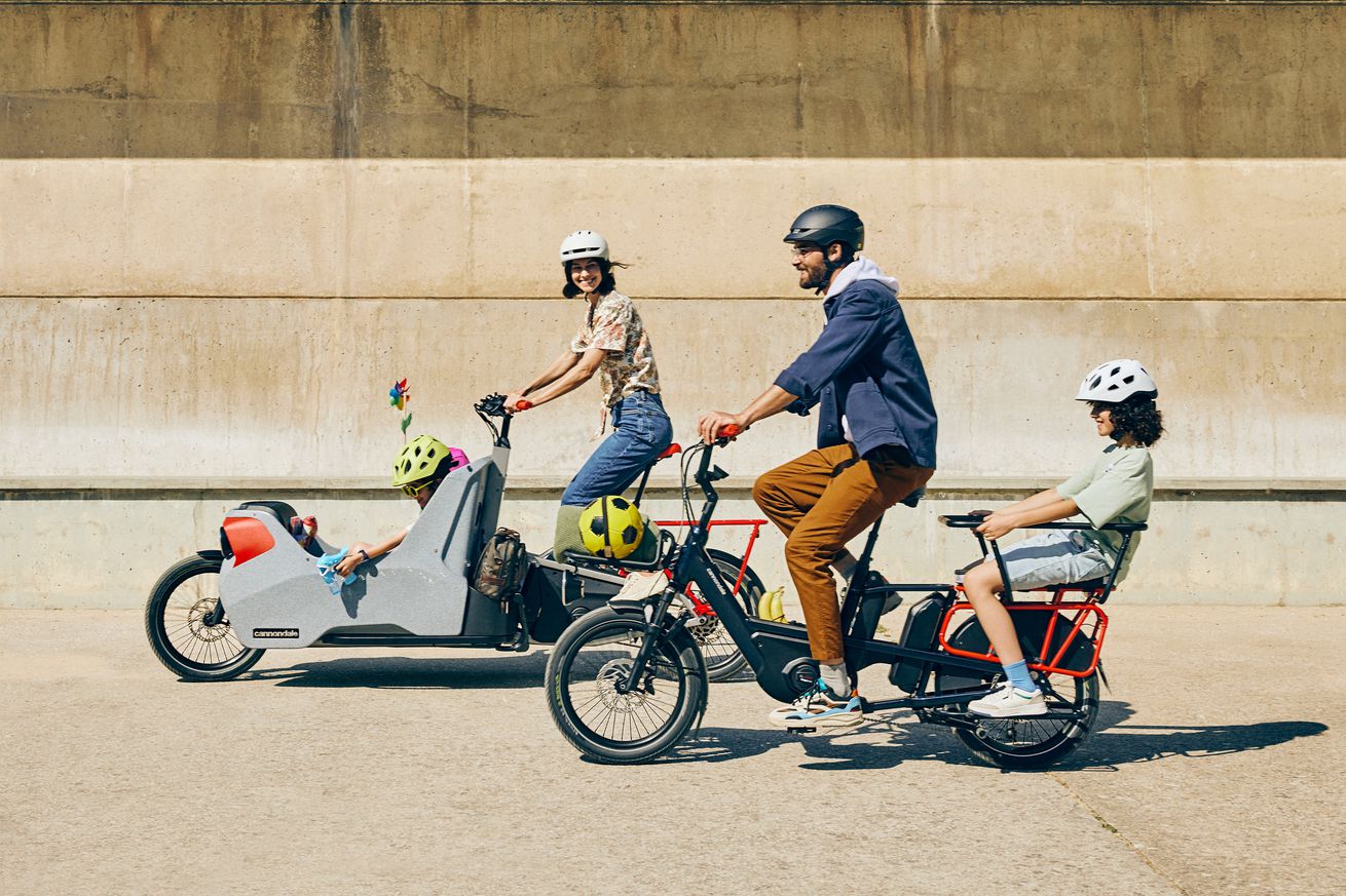 A woman rides an empty bakfiets-style Wonderwagen Neo e-cargo bike next to a man riding a Cargowagen Neo long-tail e-cargo bike with a child on the back. Everyone is wearing helmets.