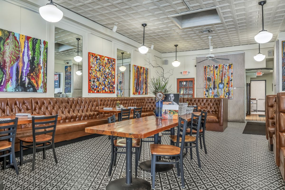 A restaurant interior with textured tile floors, loud colorful canvasses on the walls, and leather banquettes 