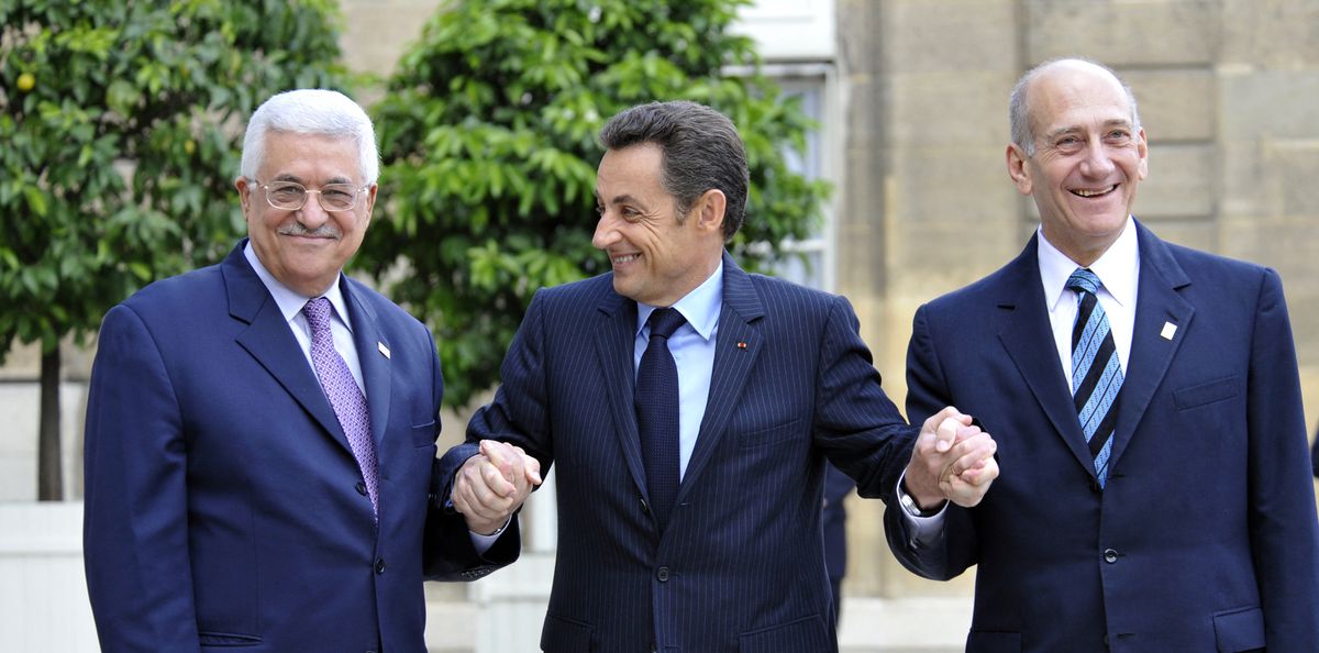 Sarkozy, in the center, grins at Abbas while holding both men’s hands; Abbas smiles, his lips pressed together, while Olmert gives a broad smile. All three men wear blue suits; Sarkozy and Olmert blue ties; Abbas, a purple tie. They stand in a courtyard, stone buildings and green trees behind them.