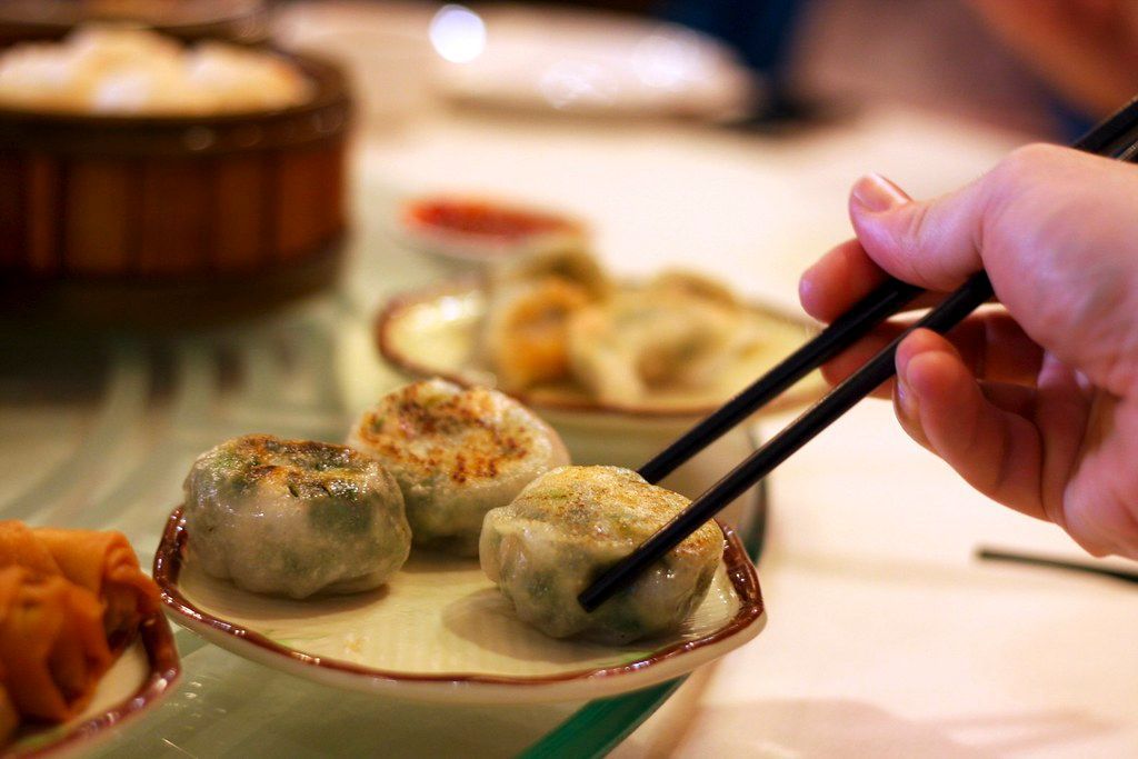 A person using chopsticks to grab one of three pan-fried chive dumplings on a plate. 