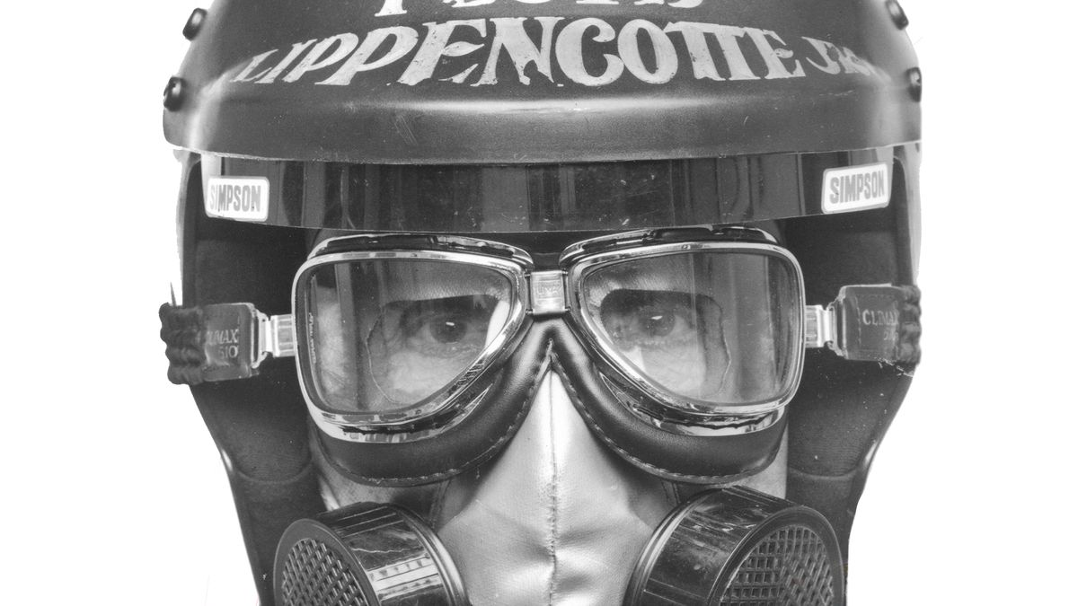 A close up black and white photo of Bob Muravez’s face as Floyd Lippencott, wearing a racing helmet and facemask, staring straight into the camera.
