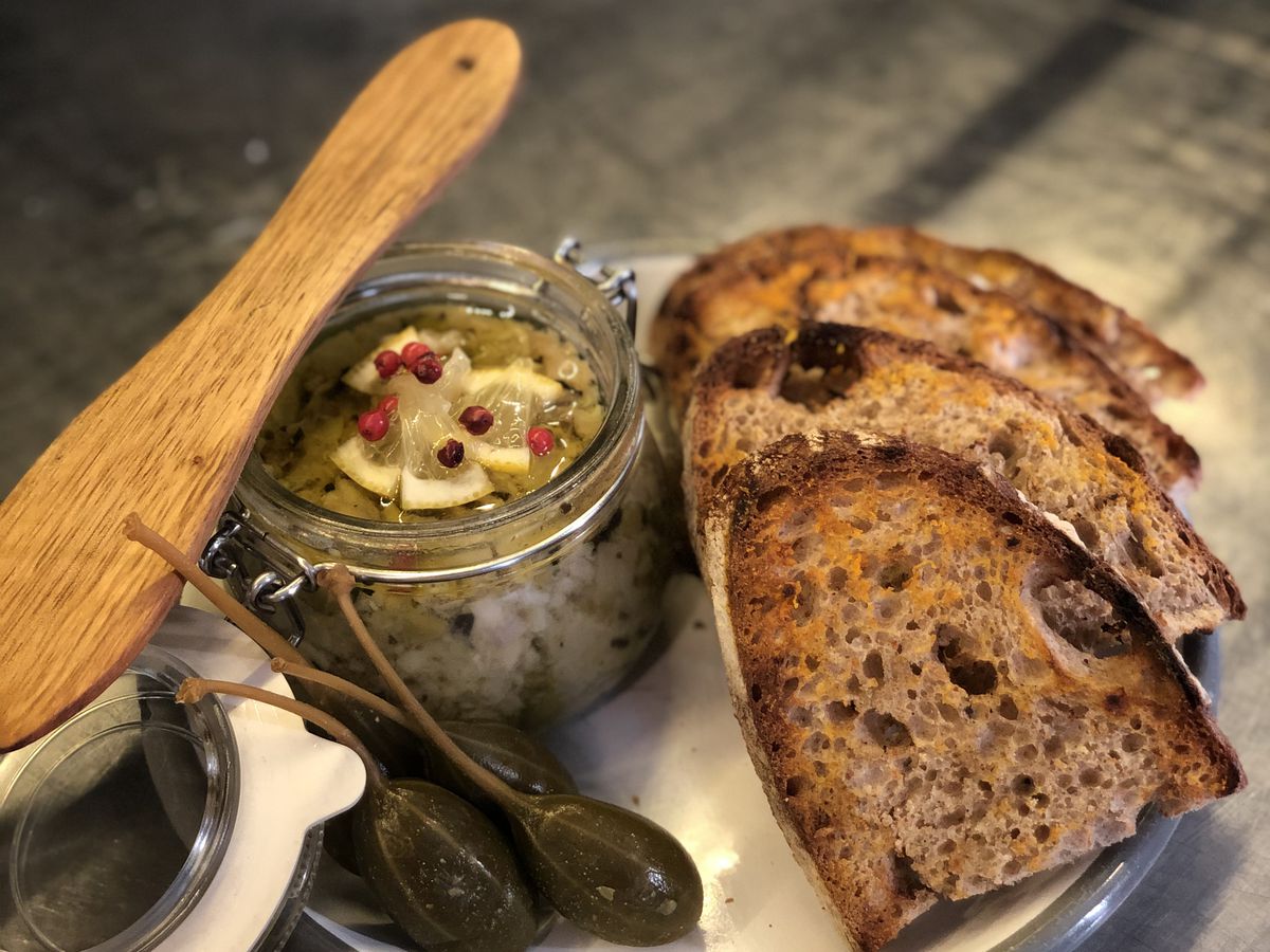 A swing-top jar with chunky spread and wooden serving spoon beside slices of toasted bread and pickled vegetables