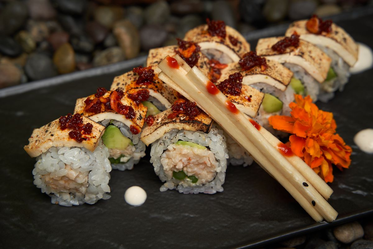 A slate plate holds rolls of sushi with vegan ingredients including blackened tofu on top.