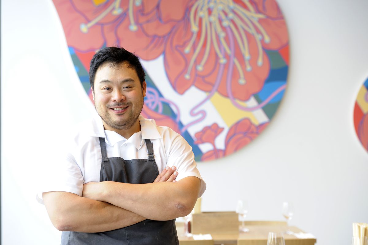 Smiling David Chang, dressed in a short sleeve white button up and dark gray apron, stands in front of a colorful, abstract wall mural with his arms crossed.