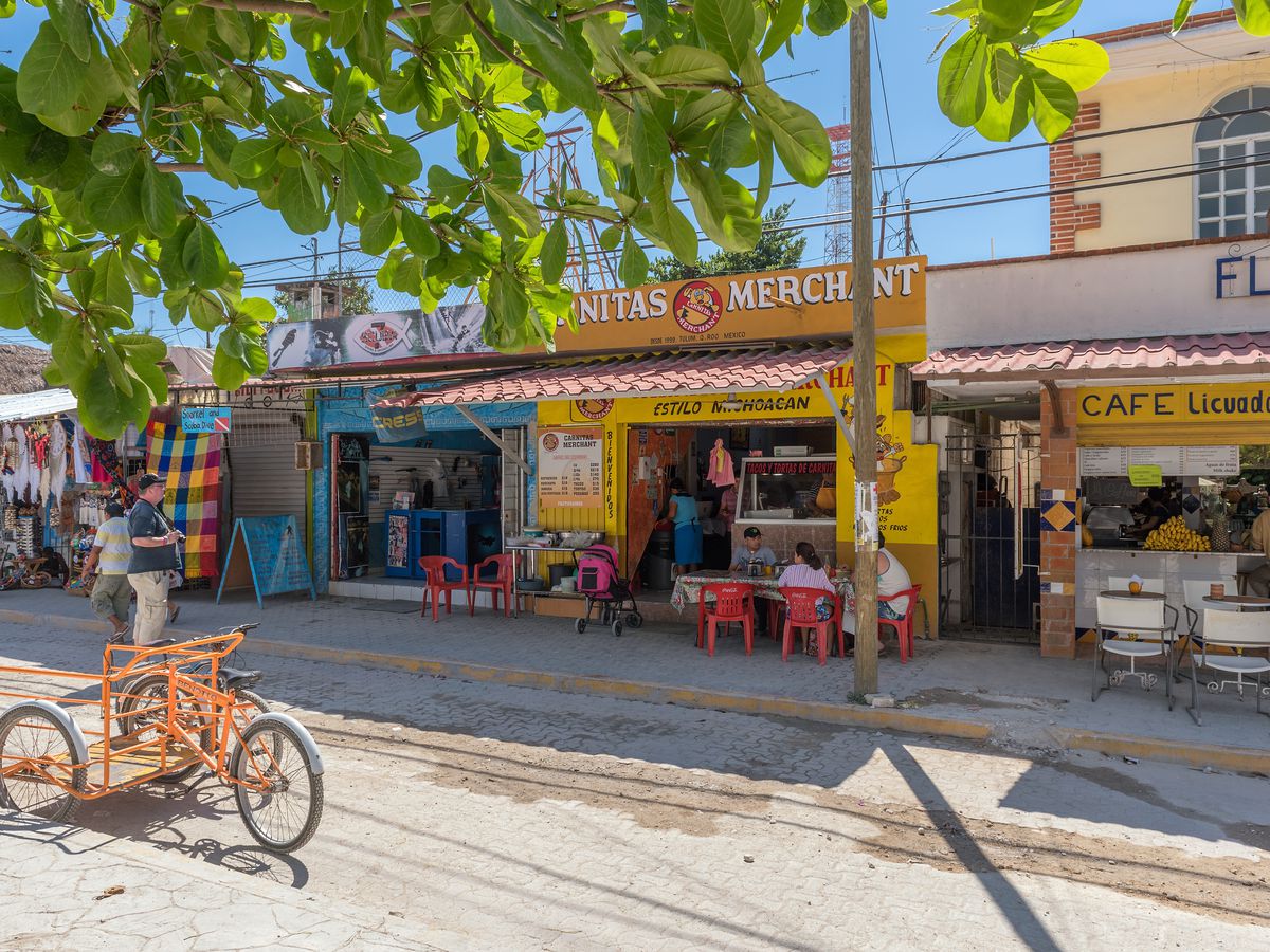 A row of shops and restaurants, with a few customers sitting at sidewalk tables, on a sunny street in Tulum. 