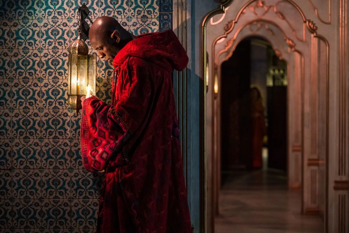Idris Elba, in a red robe, stands in front of a brilliantly-patterned wallpaper.