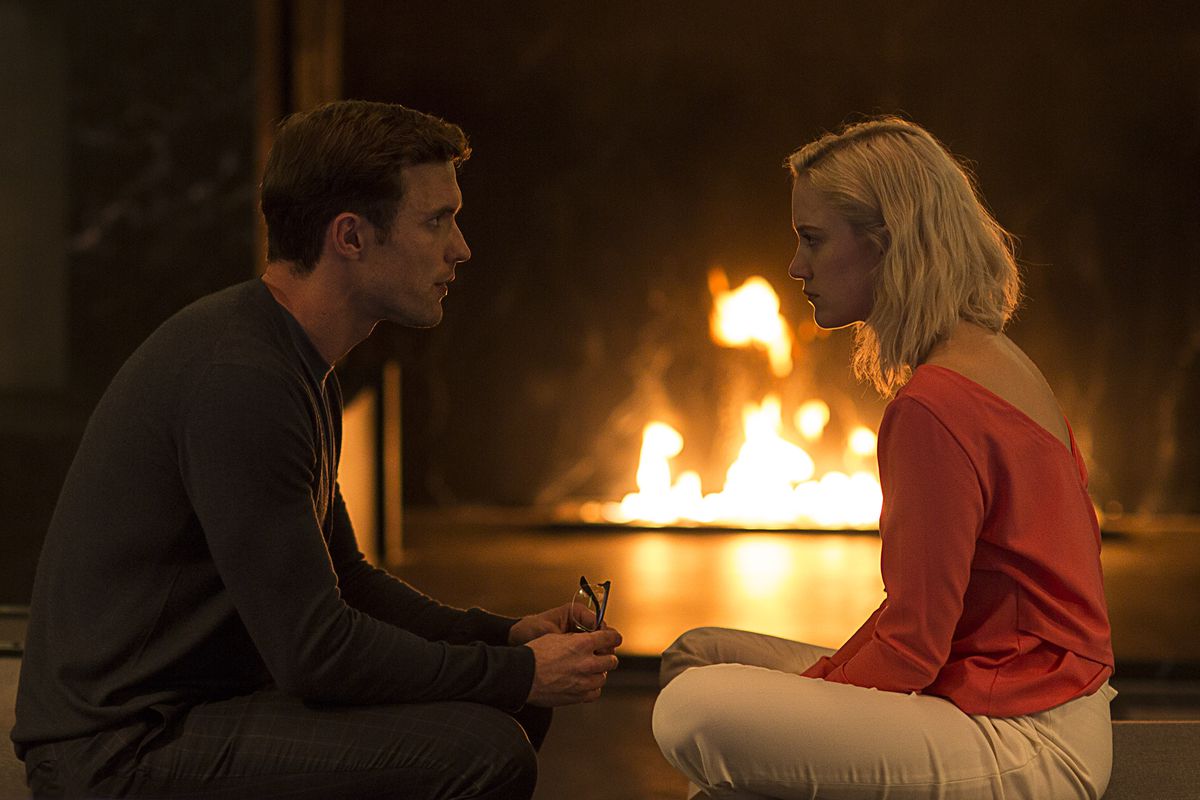 Ed Skrein and Maika Monroe sit next to each other with their legs folded in Tau. A fireplace burns in the background, and Skrein holds a pair of glasses in his hands. They look at each other.