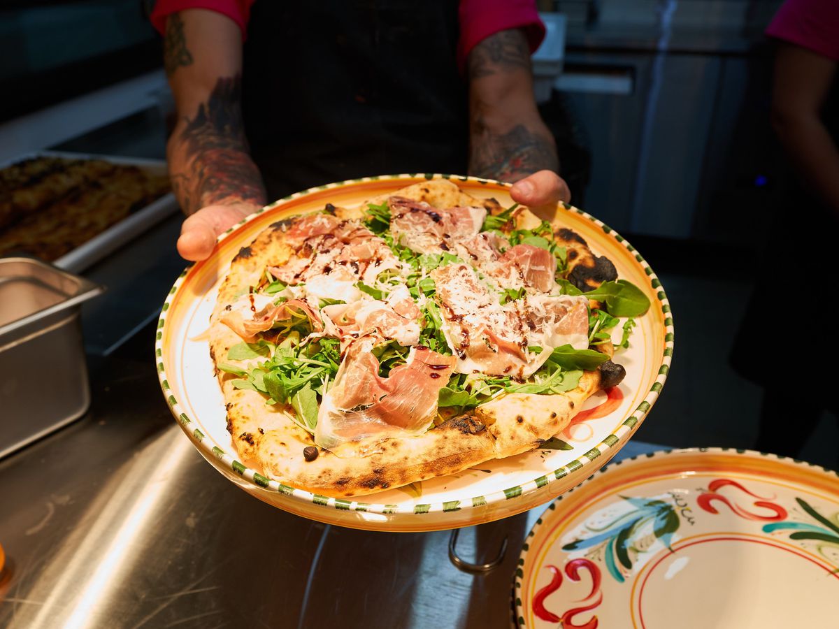 A pair of hands holds a whole pizza topped with thin ribbons of prosciutto and fresh arugula