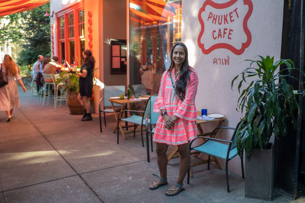 A woman in a pink dress stands outside a restaurant with the words “Phuket Cafe” painted on the pink exterior 