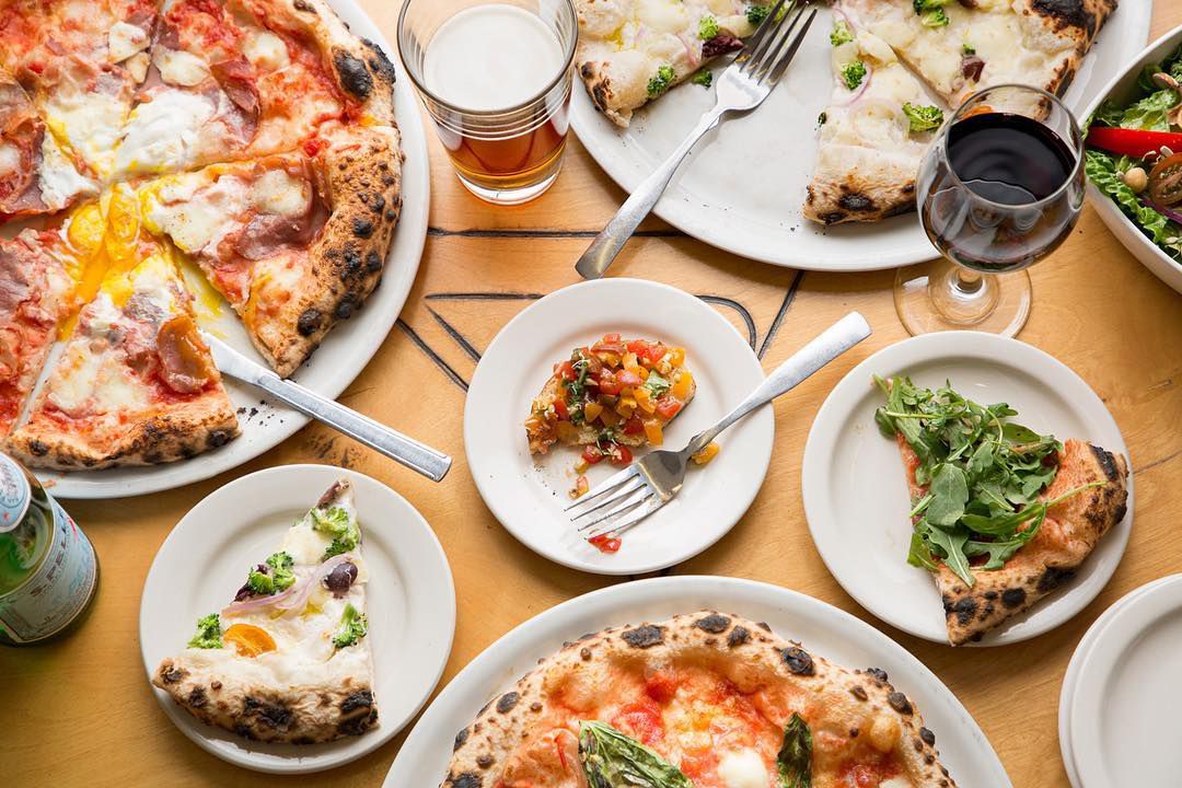 From above, a table filled with various pizzas in different states of deconstruction, a few small plates with individual slices, utensils and glasses of beer