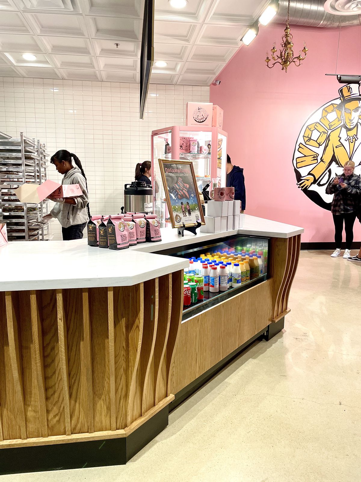 In a doughnut shop with a pink wall and white subway tiles, a woman stands at a rack and loads goods into a pink bakery box. 