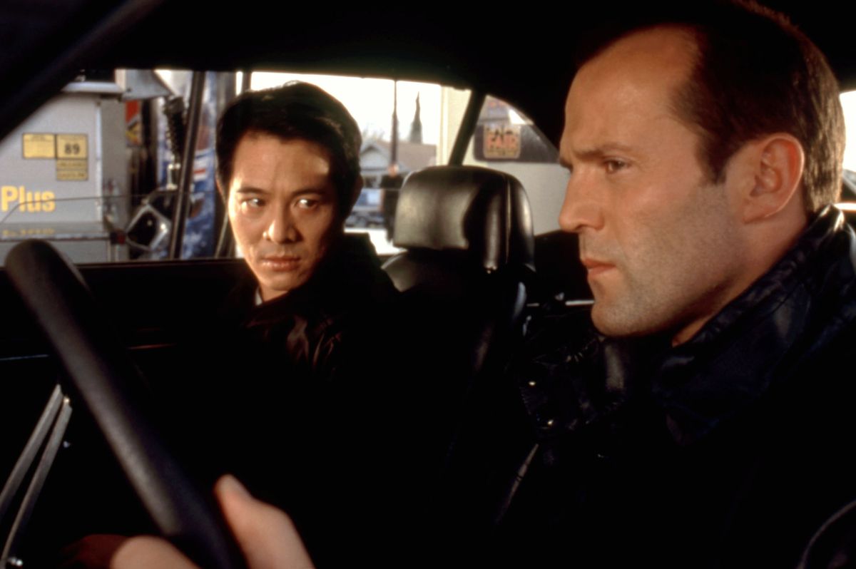 Jet Li and Jason Statham sit in a car together. Li, sitting in the passenger seat, looks at Statham (who has hair), who is in the driver’s seat, in The One.