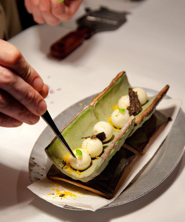 A chef adds the finishing touch to a row of meringue like puffs in a long vegetale-looking tube dotted with various colorful garnishes on a long ceramic plate on a prep station